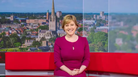 BBC Susie Fowler-Watt, wearing a cerise-coloured dress, sits on the red Look East sofa. She is smiling with her hands clasped. The studio backdrop shows a view of Norwich with landmarks including both cathedrals and City Hall 