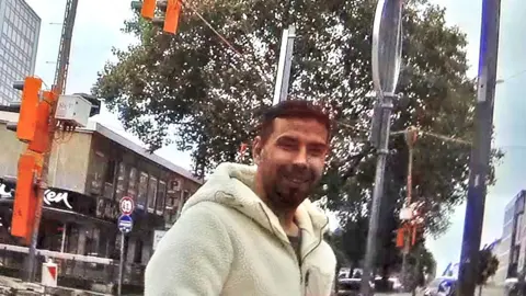 A dark-haired man with a beard and white fleece jacket smiles unsuspectingly at our reporter. He is standing outdoors with a parade of shops, a tree and street furniture surrounding him.