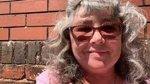 Rochelle Russell Rochelle Russell with curly hair and glasses against a red brick wall