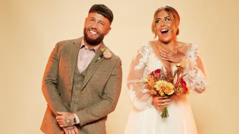 Channel 4 Polly and Adam in wedding outfits, with her wearing a white dress with sheer arms and a low v-neck and holding a bouquet of orange and red flowers while Adam is wearing a tweed suit in a pale brown with a pastel pink shirt and buttonhole.