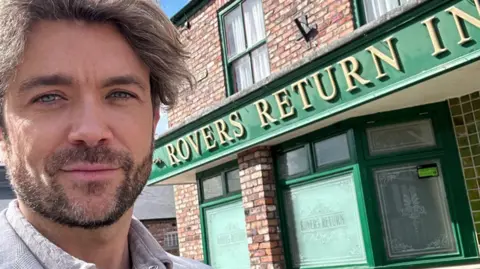 Emrhys Cooper Emrhys Coopers stands in front of Coronation Street's Rovers Return Inn. The pub has a red brick exterior with a green facade. The letters 'Rovers Return Inn' are written in gold.