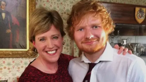 Susie Fowler-Watt/BBC Susie Fowler-Watt, wearing a maroon and black coloured dress, pictured with Ed Sheeran, who is wearing a white shirt and maroon tie