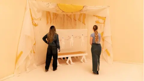 Josh Croll/Tate Photography Two women stand with their backs to the camera looking at a fabric installation painted with yellow hands and feet, and a painting sitting on a white table held up by designs of human legs and feet.  