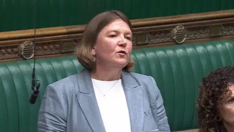 BBC A woman speaking in front of a green bench, with a microphone to her side