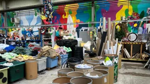 The inside of Scrapstore. There are large bins and barrels filled with craft supplies including felt, fabric, cardboard, scraps, paints, stencils, wood and more. The walls of the warehouse are painted bright colours in a wiggly pattern.