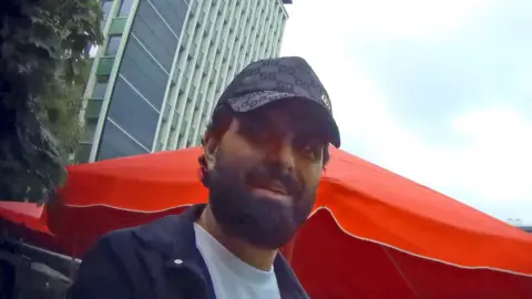 A dark-haired bearded man wearing a Gucci baseball cap, white t shirt and navy shirt, looks at the undercover reporter. An orange cafe umbrella and a high-rise building are seen in the background.