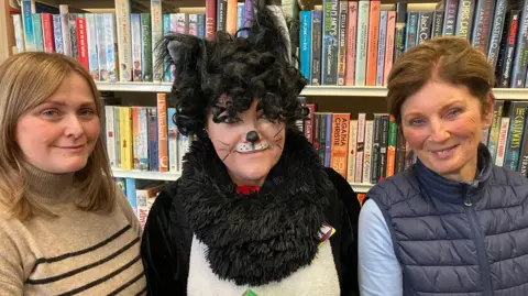 BBC Vicky Fedak who has long light brown hair, wearing a beige and black striped top with the Library Cat who is dressed up as a cat with black and white fur and her face is painted with black whiskers and nose. Carol is standing to the right and has brown hair that's tied back, she's wearing a navy gilet and blue top. They are all smiling and standing in front of a bookcase full of books. 