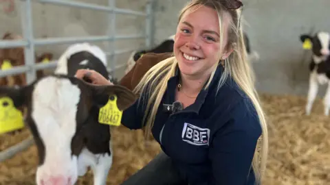 BBC A young blonde woman smiling at the camera and stroking a brown and white calf