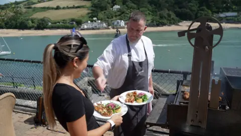 BBC A chef serves food from a BBQ on a restaurant terrace by a river