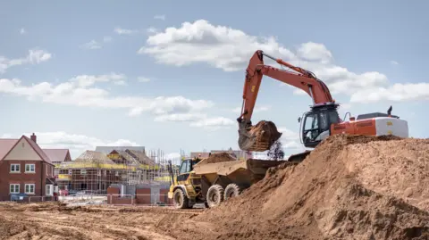 Getty Images A stock image of a construction site. A digger can be seen on top of a large pile of earth, lifting it into the back of a truck. In the background there are new-build houses, some of them covered in scaffolding. 