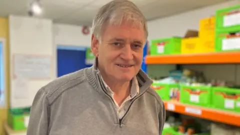BBC Phil Stratton has a grey zip-up jumper and a white shirt. He is white and has white hair