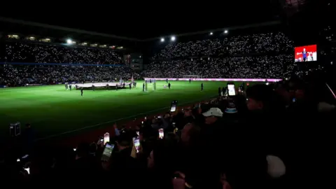 CUE Audio A football ground at night, with the pitch floodlit and many lights visible in the stands from mobile phones being held up by fans