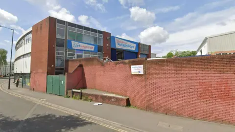 Google The outside of the Scrapstore building. It is a large brick warehouse with lots of windows on one side and white cladding on the other. It has a faded sign on the front which says 'Children's Scrapstore'. There is a long red brick wall which encloses the car park, directly outside the entrance to the shop.
