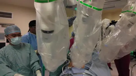 An operation being carried out in an operating theatre. Robotic arms covered in plastic hold the instruments over the patient's body. A surgeon in medical dress is standing beside them.