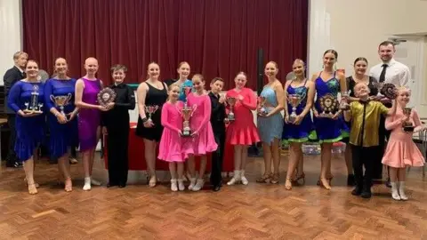 Southend Dance Centre Various ballroom dancers in bright outfits holding trophies.