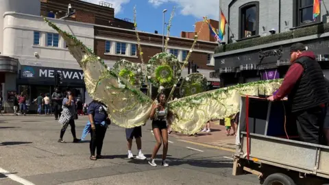 BBC/Peter Cooper Carnival parade with woman wearing huge yellow wings in the foreground