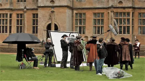 BBC A group of actors dressed in Tudor clothing standing the the grass outside Montacue House, which is a large Elizabethan mansion made from stone. There is a film crew holding microphones above the actors and a large light reflector sheet is positioned behind the camera.