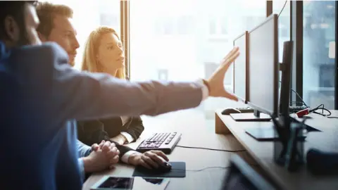 Getty Images Two men and a women gathered around a dual screen in an office.