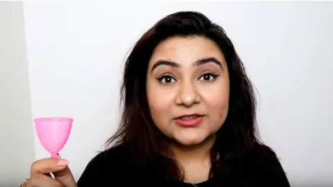 The Delhi Fashion Blogger  Komal holds up a pink plastic menstrual cup in a YouTube grab