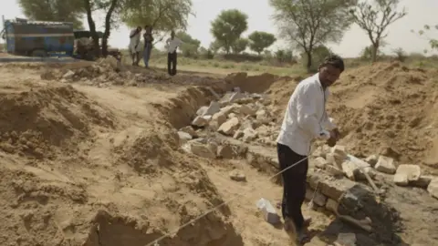 Man building a check dam, measuring the landscape with rocks around him while three other men watch