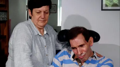PA Jane stands next to her husband, who is using a wheelchair and is wearing stripy shirt. 