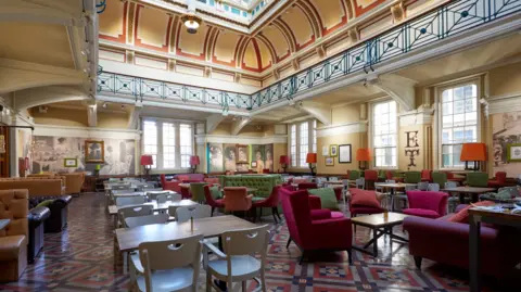 David Rowan Chairs and tables fill a bright, spacious room, the walls are cream, except for the back wall which has portraits and photographs covering it