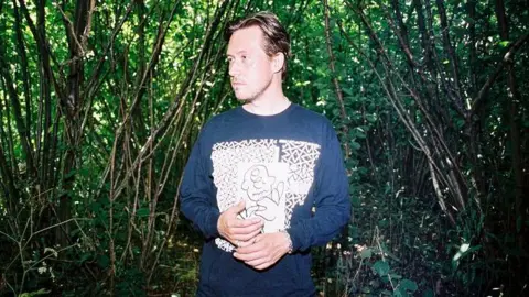 Emma Toma DJ Joy Orbison standing against a woodland backdrop. He is wearing a blue and white sweatshirt