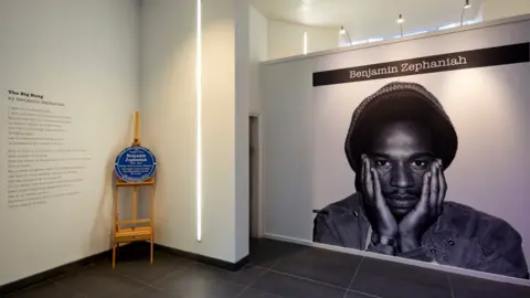 David Rowan A black and white photo of Benjamin Zephaniah, which fills an entire wall, is next to a blue plaque resting on a wooden stand