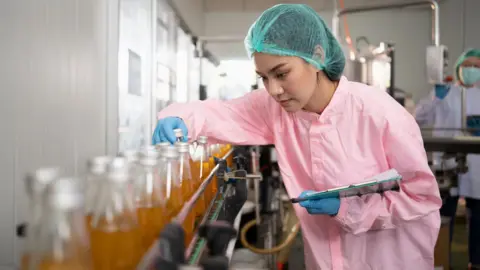 Getty Images A woman in a blue hair net wearing a pink coat and blue gloves inspects a glass bottle on a factory production line