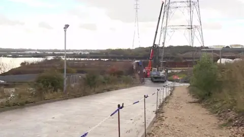 BBC View of the entrance to the Howdon treatment works, with the banks of the River Tyne to the left, a road with a digger at the end of it, next the the lower part of an electricity pylon.