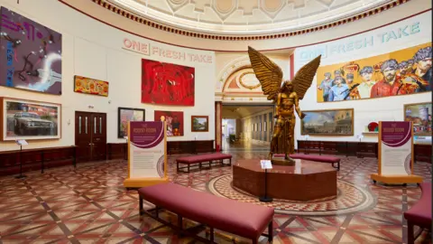 David Rowan A circular room with a golden statue of an angel in the centre, it is surrounded by red benches and has two floor-standing signs either side of it. On the walls are multiple pieces of art, and at the top of the walls on both sides of the image are the words "One Fresh Take"