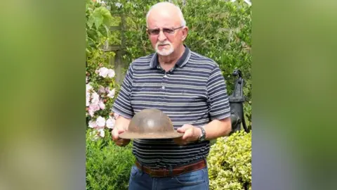 Le Paradis Massacre website Dennis O'Callaghan, with white hair, beard and glasses, wearing a blue striped short-sleeved shirt while stood in a garden. He is holding a World War One British helmet, which was refurbished in the late 1930s and used during World War Two.