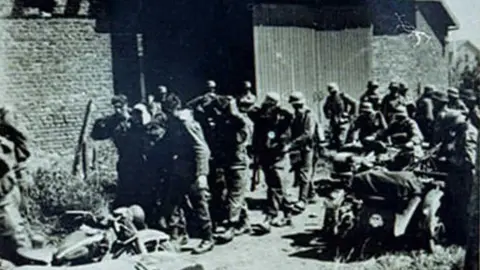 Dennis O'Callaghan A blurry black and white photo taken in 1940 showing British soldiers surrendering to SS soldiers. There is a barn behind them and two of the British soldiers are holding up a third. A much larger group of SS soldiers are surrounding them, holding guns or sitting on their motorcycles.