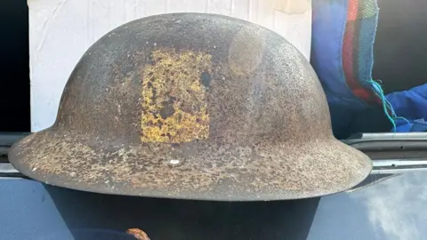 Le Paradis Massacre website A World War One British infantry helmet repurposed for World War Two. It has a rivet on its rim where a new chin strap and lining would have been added. It has a yellow rectangle on its side with bits of black pigment.