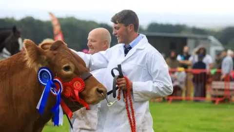 CALLUM STALEY/CSJ PHOTOGRAPHY A young man wearing a white coat holds the halter of a brown cow. The halter stap has four blue, white and red rosettes attached to it and there is an older man, also wearing a white coat, smiling in the background.