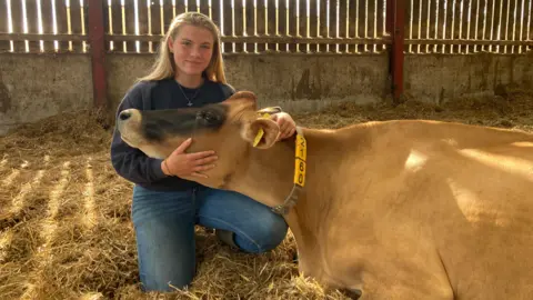 BBC Erin Gailey in a barn holding a cow's head. She has long, light hair past her shoulders and wears a blue top with a gold necklace and blue jeans as she kneels on straw in an enclosed building. The cow has brown fur apart from darker fur on its nose and a plastic necklace with numbers on it around its neck and a yellow tag in its ear
