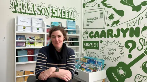 Culture Vannin  A woman sitting in a classroom with textbooks on a stand behind her, and a green mural with Manx language phrases written on the wall. 
She has brown wavy hair, and is wearing a blue sweater with white stripes. 