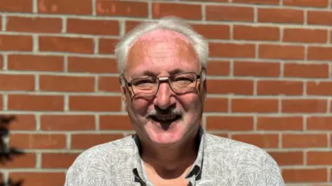 George King/BBC A man with glasses and grey hair smiling into the camera while standing in front of a brick wall.