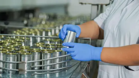 Getty Images A worker wearing blue latex gloves handles empty silver tin cans on a factory production line