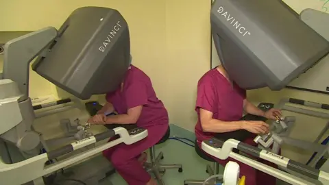 BBC Two surgeons in maroon medical dress sit back-to-back with their heads in large grey consuls with the word 'Davinci TM' on them