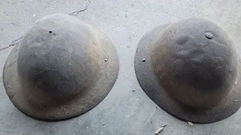 Le Paradis Massacre website Two World War One helmets repurposed for use in World War Two. They are lying rim down on the ground and are rusty.