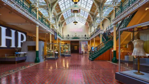 David Rowan A glass-roofed building contains items on display in cabinets on left and right sides with a long red floor and HP sign on one side