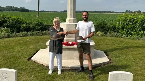 Le Paradis Massacre website Biddy Fisher wearing a grey top and white trousers shaking hands with Jean-Charles Dufour at the white stone Le Paradis war memorial. She is holding one helmet while he is holding another. Behind them are green fields and sky.