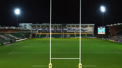 Getty Images Franklin's Gardens rugby ground at night with goalposts in the foreground and stands on three sides. Two floodlights are visible left and right, and a large display screen is seen to the right.