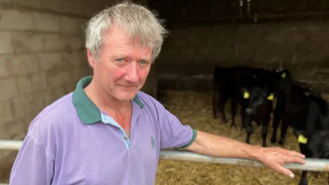 BBC A man wearing a purple polo shirt with green edging stands by a barn with hay on the floor and cows behind him while he leans his left arm on the metal gate