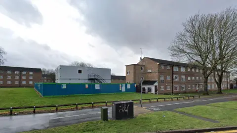 Google Brown brick blocks of flats surround a green space, on which sits a grey temporary building, surrounded by blue fencing.