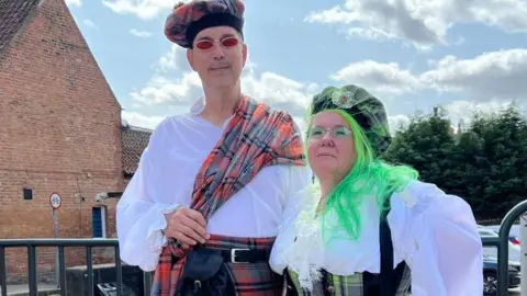 Amber Macey/BBC Man with red kilt and white shirt wearing a tartan beret next to woman with green kilt wearing a white lacey shirt with bright green hair
