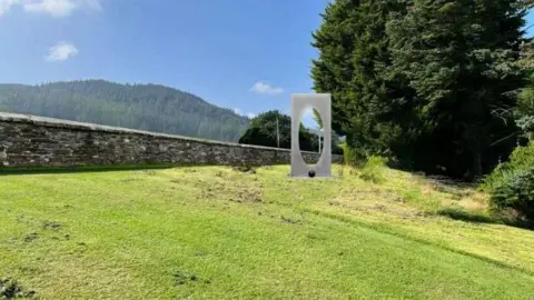 Albany Arts Communications A steel modern art sculpture, which is a rectangle with an oval shaped hole, standing on some grass. It is placed next to a low stone wall, with a tree-covered hill in the background.  