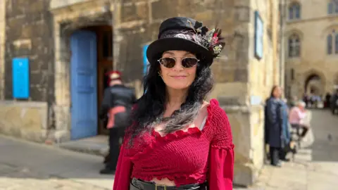 Amber Macey/BBC Woman dressed in red top with dark sunglasses and top hat decorated with feathers