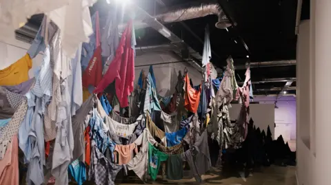 Blake Watson Photography Shirts of different colours, designs and sizes are hung indoors around lines that drop down close to the floor where some of the shirts are touching the floor. There are hundreds of shirts on display.